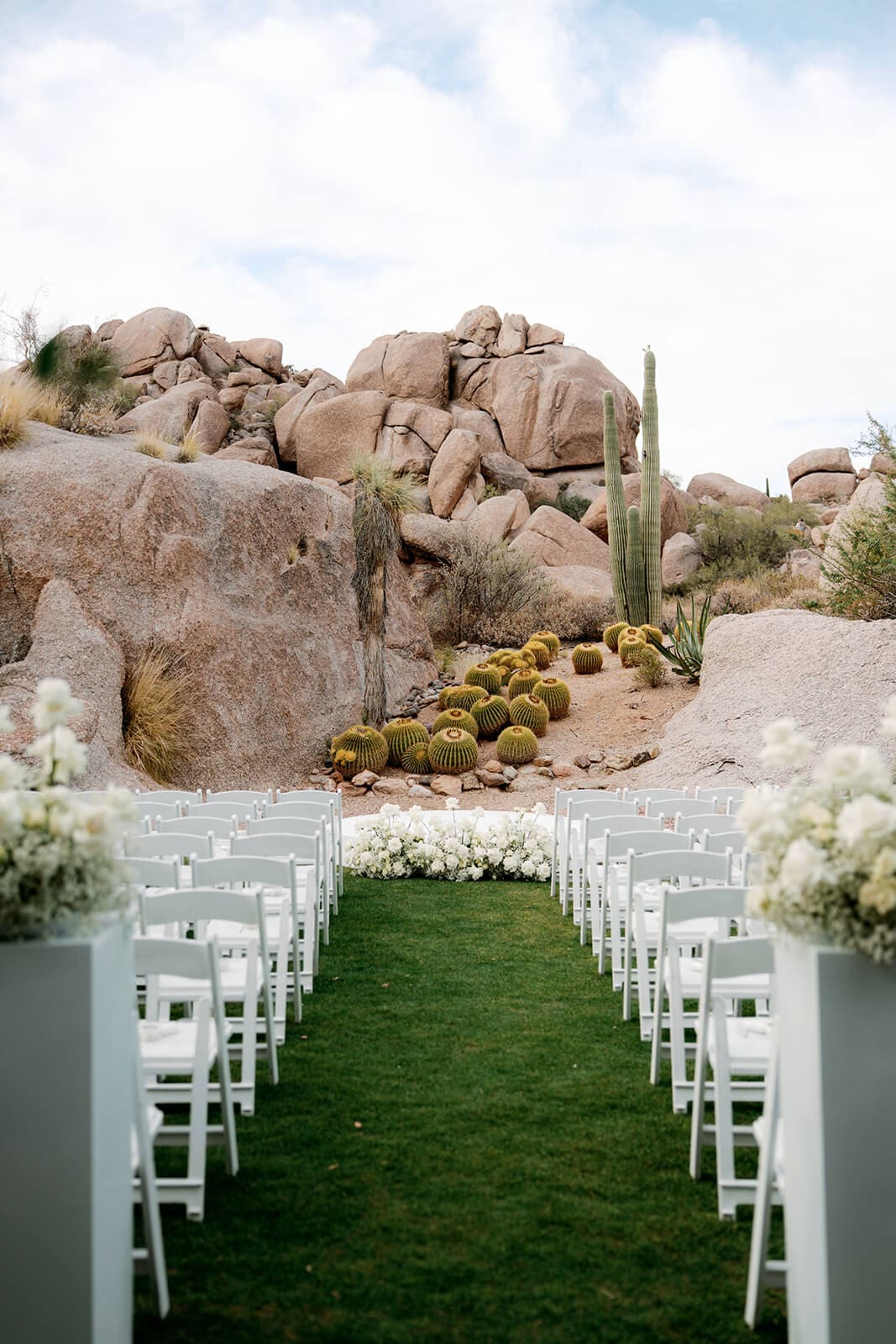 Outdoor wedding ceremony aisle lined with white chairs and floral arrangements overlooking saguaros, barrel cacti, and desert boulders.