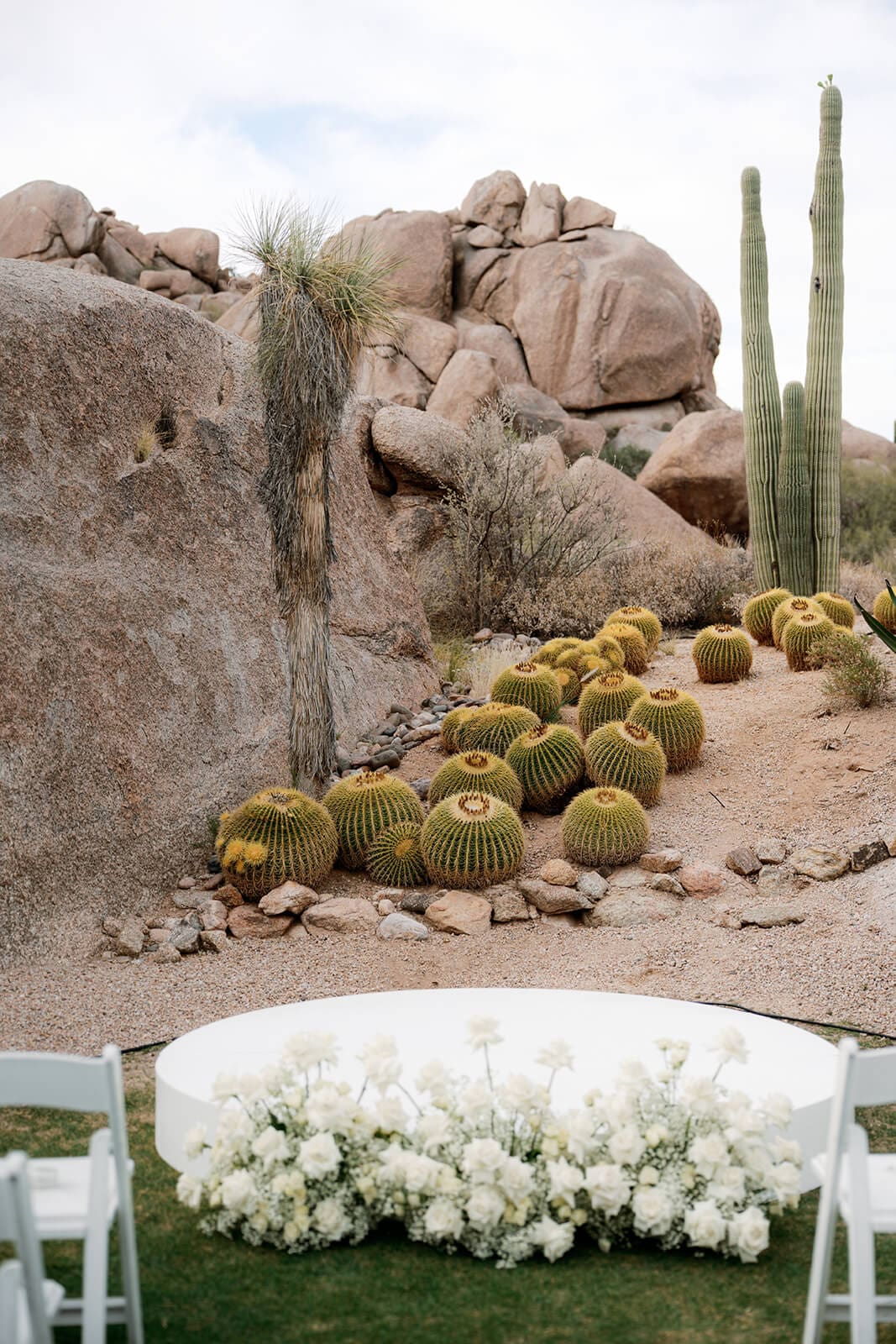 Outdoor desert wedding ceremony setup with white flowers, barrel cacti, tall saguaros, and large granite boulders.