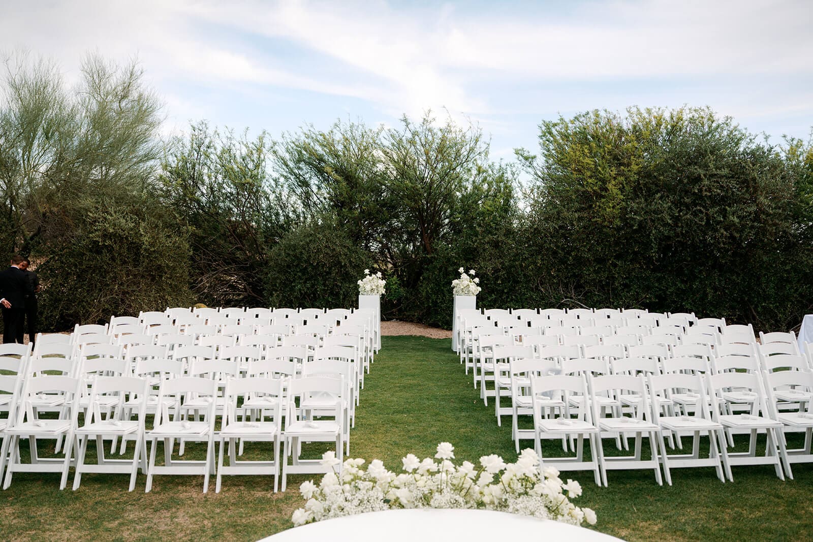 Rows of white ceremony chairs arranged on a lawn with white floral arrangements and dense desert greenery surrounding the outdoor wedding space.
