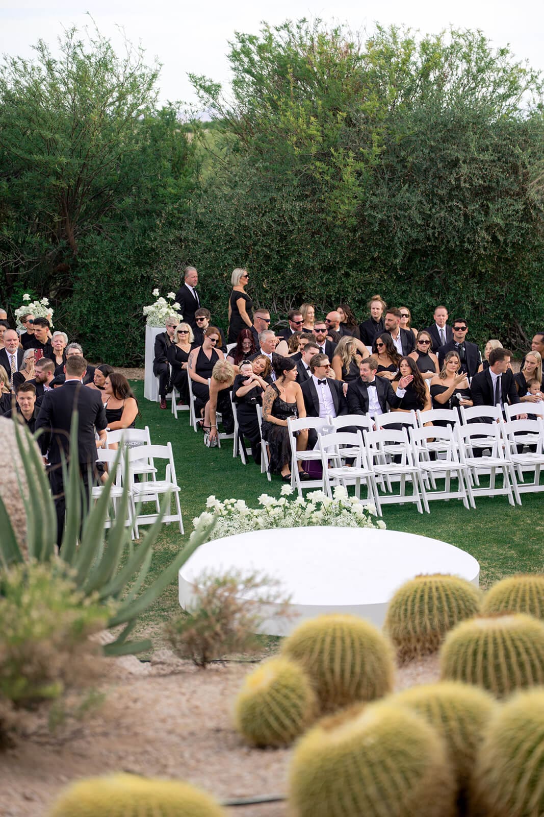 Wedding guests seated in white chairs during an outdoor desert ceremony surrounded by greenery, cacti, and granite rocks.