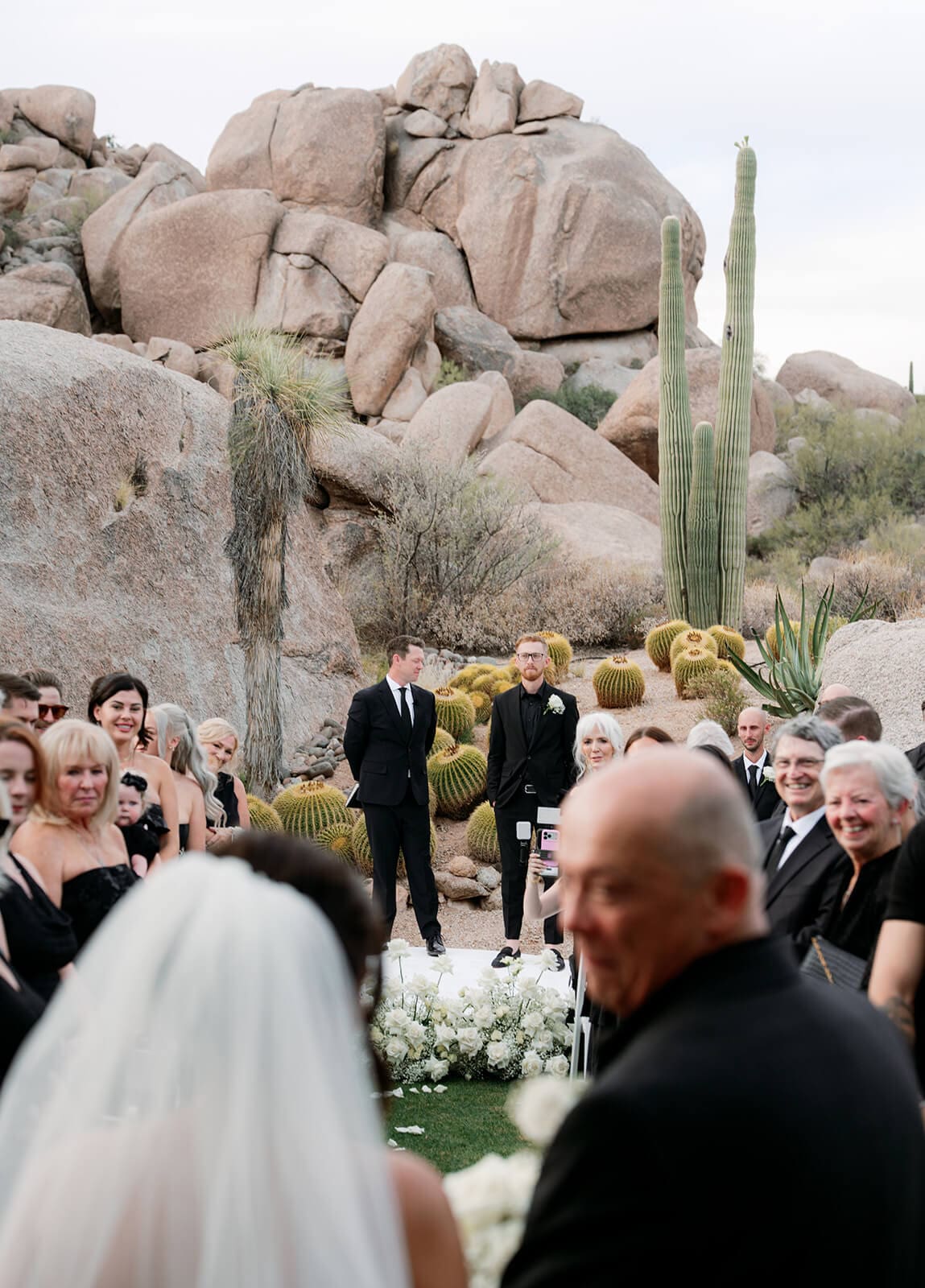 Bride walking down an outdoor wedding aisle toward the groom with large desert boulders, saguaros, and barrel cacti behind the ceremony.