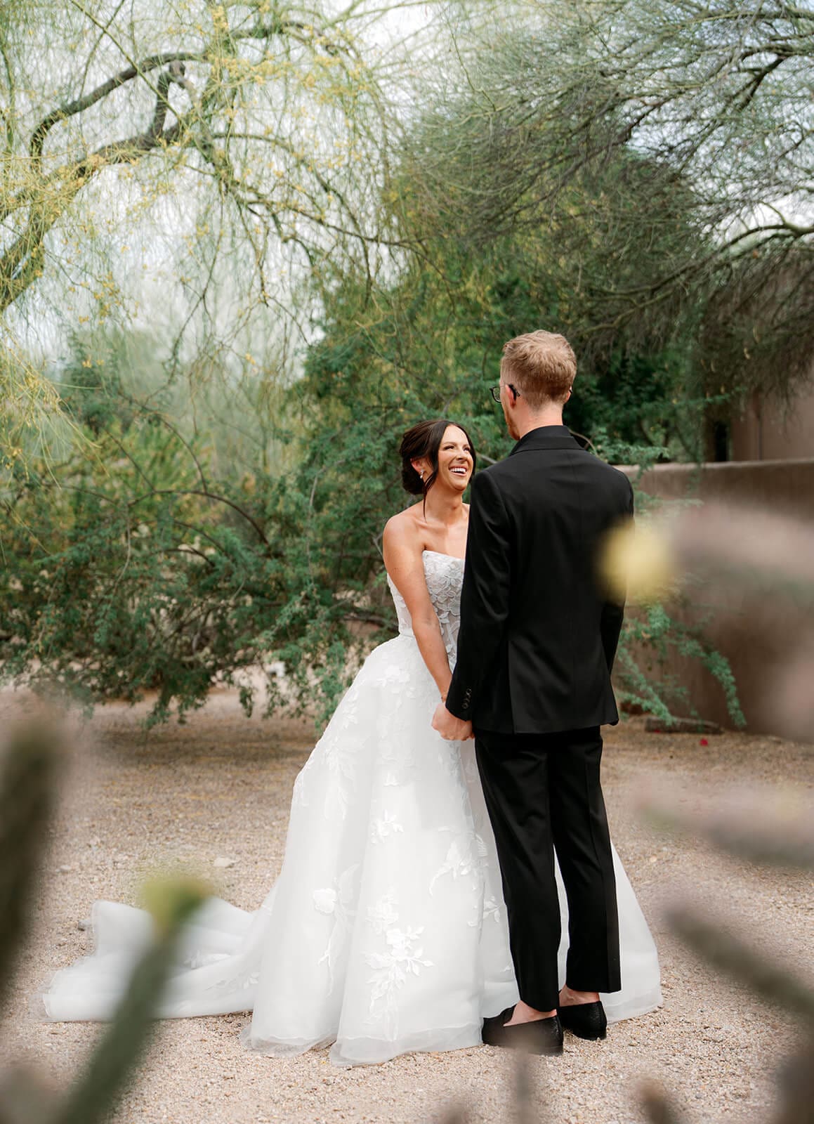 Bride and groom holding hands and smiling during an outdoor first look surrounded by desert trees.