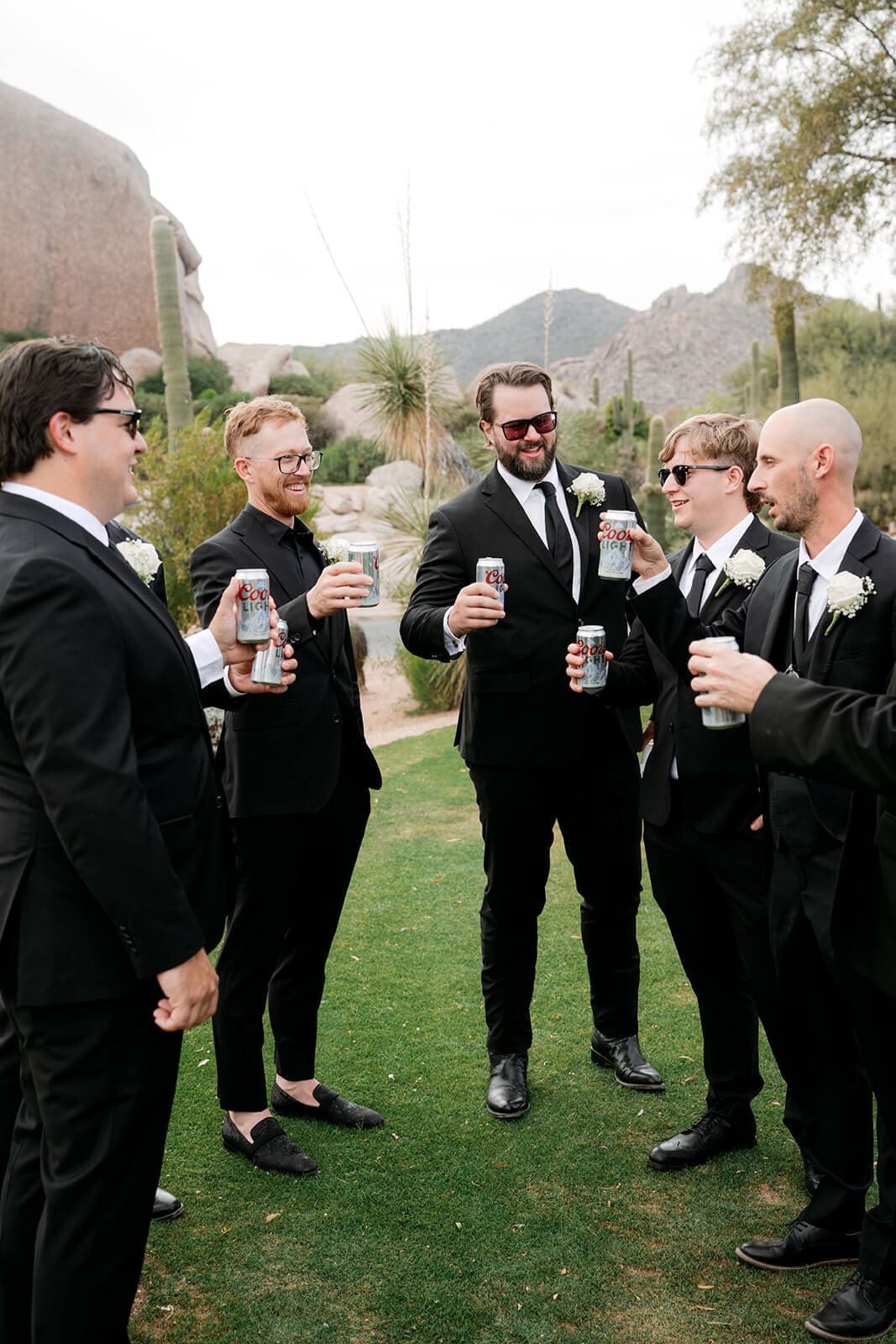 Groom and groomsmen in black suits raising cans for a toast outdoors with desert cacti and rocky mountains behind them.