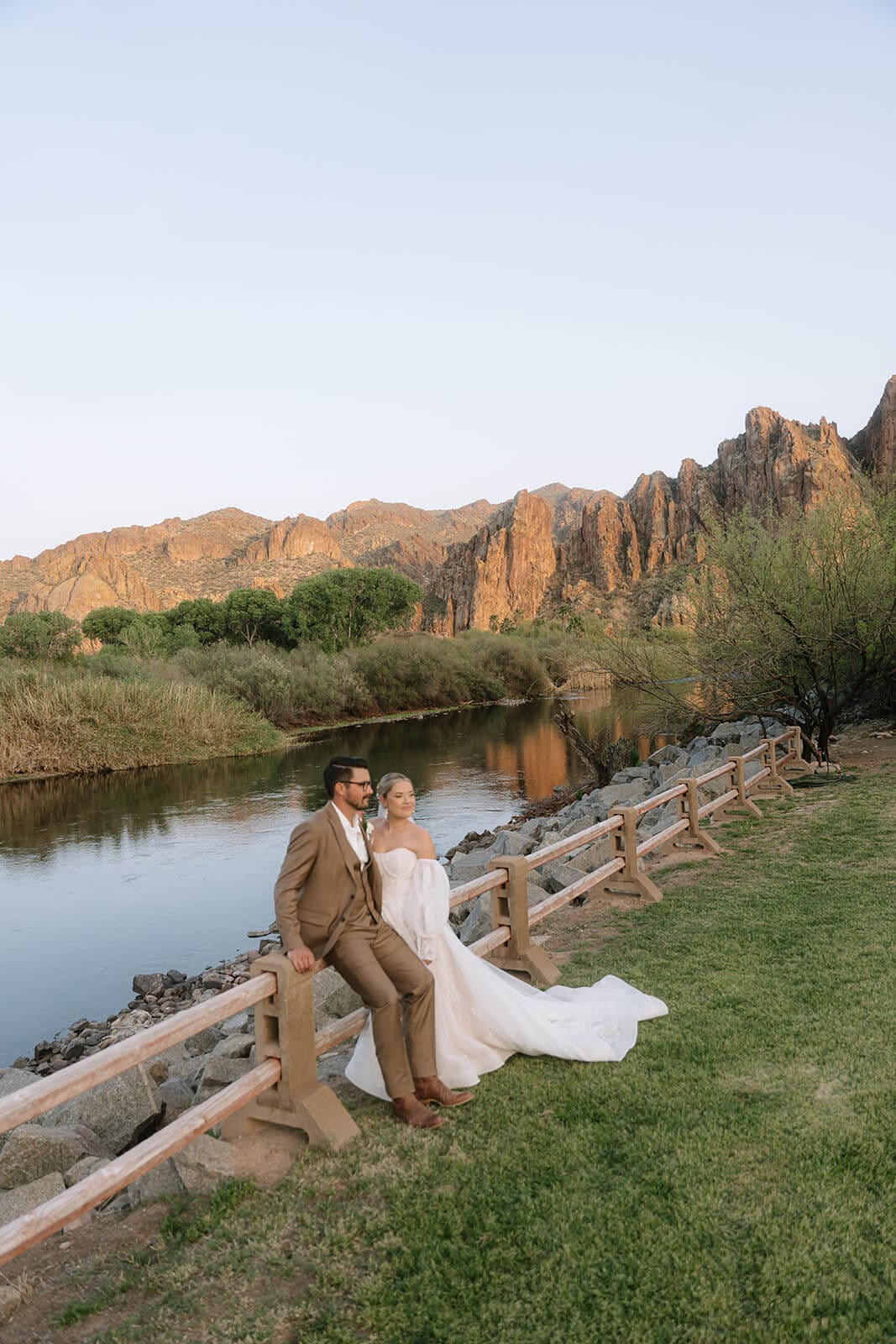 Bride and groom sit together beside a peaceful desert pond with rocky Arizona mountains glowing in the distance.