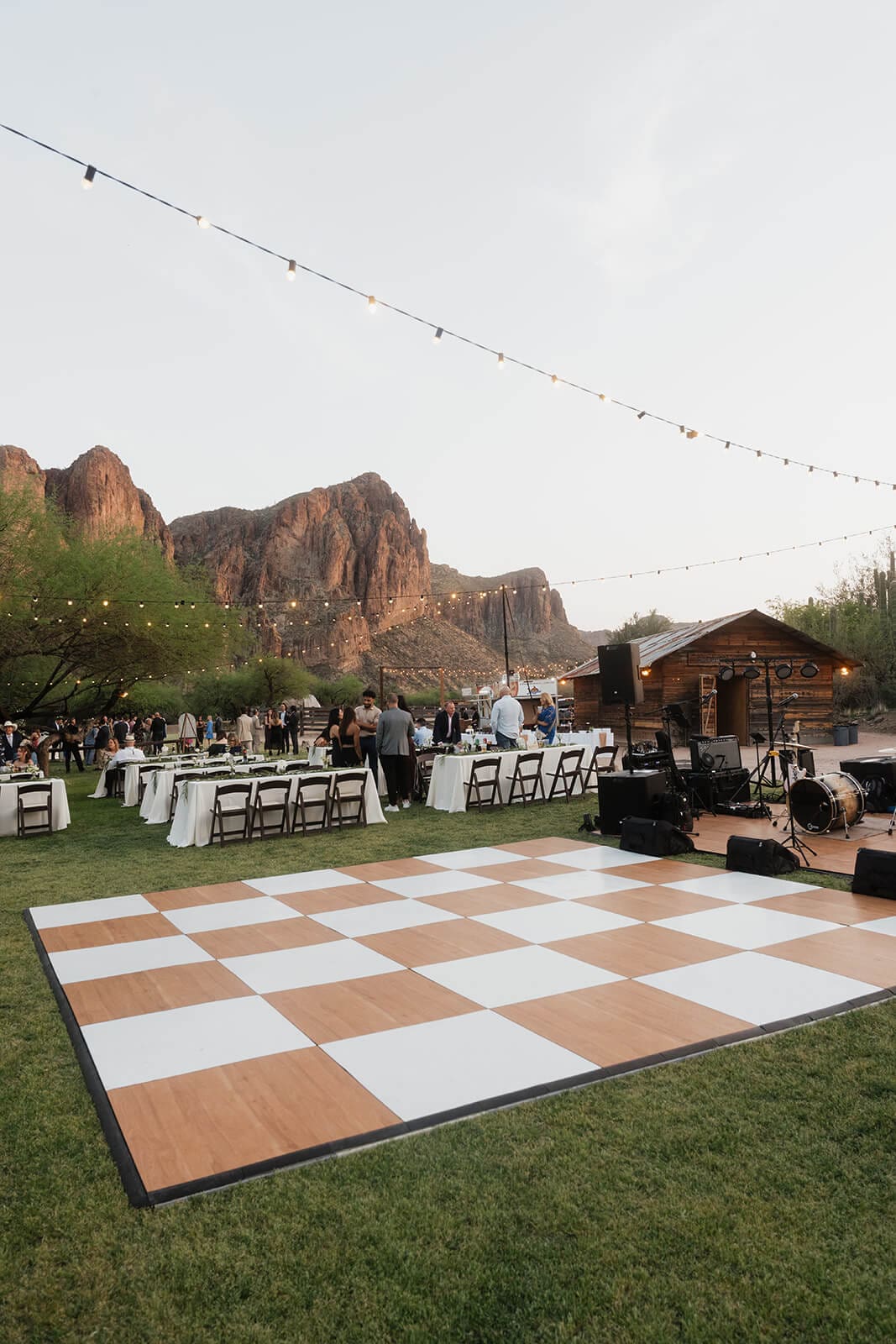 Outdoor wedding reception with a checkered dance floor, string lights, dinner tables, and the Superstition Mountains at sunset.