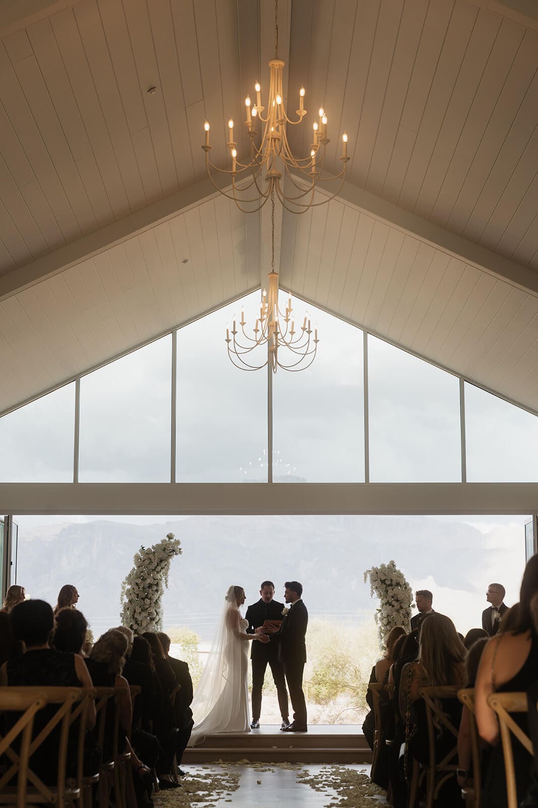 Indoor wedding ceremony beneath chandeliers and vaulted ceilings with large windows framing the desert landscape.