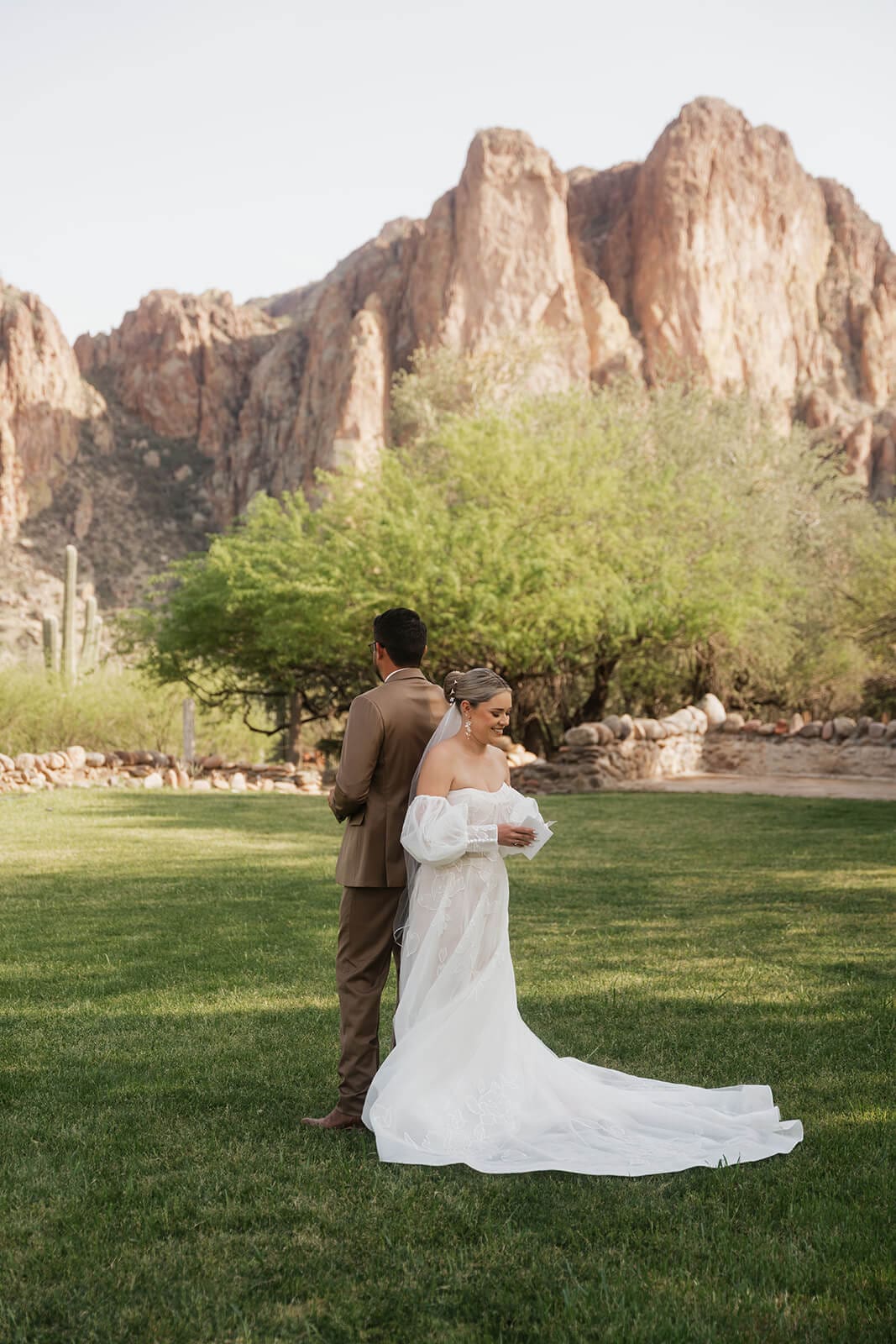 Bride and groom embrace on a green lawn beneath dramatic views of the Superstition Mountains.