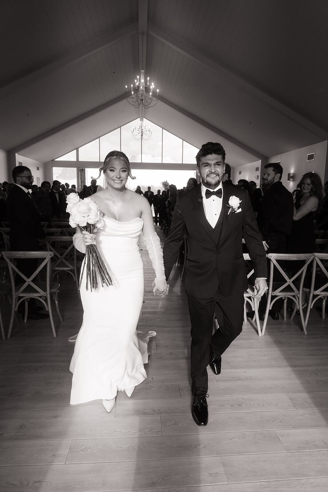 Black-and-white photo of the bride and groom smiling as they walk back down the aisle after their indoor wedding ceremony.