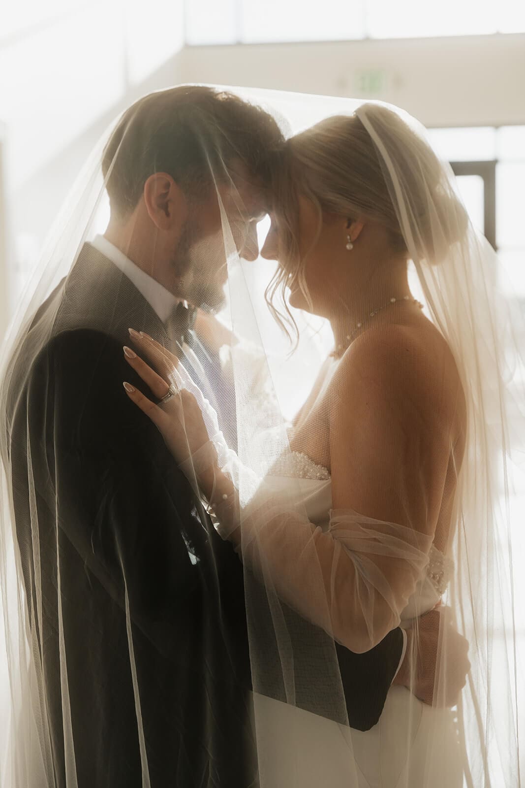 Bride and groom lean forehead to forehead in a softly backlit wedding portrait beside a bright venue window.