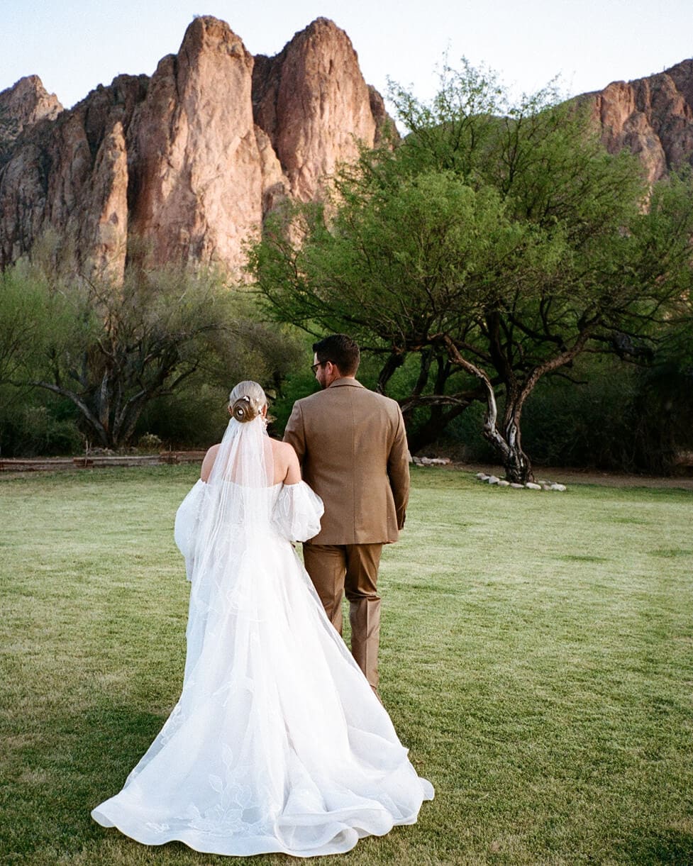 Bride and groom walk across a grassy lawn with the Superstition Mountains rising behind them at their Arizona wedding.