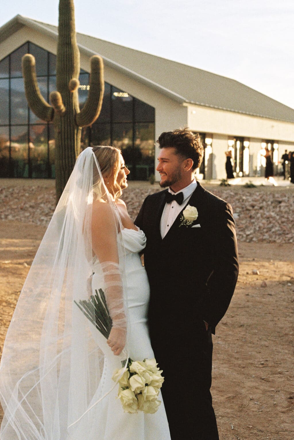 Bride and groom stand together outside a modern Arizona wedding venue with a tall saguaro cactus beside them.