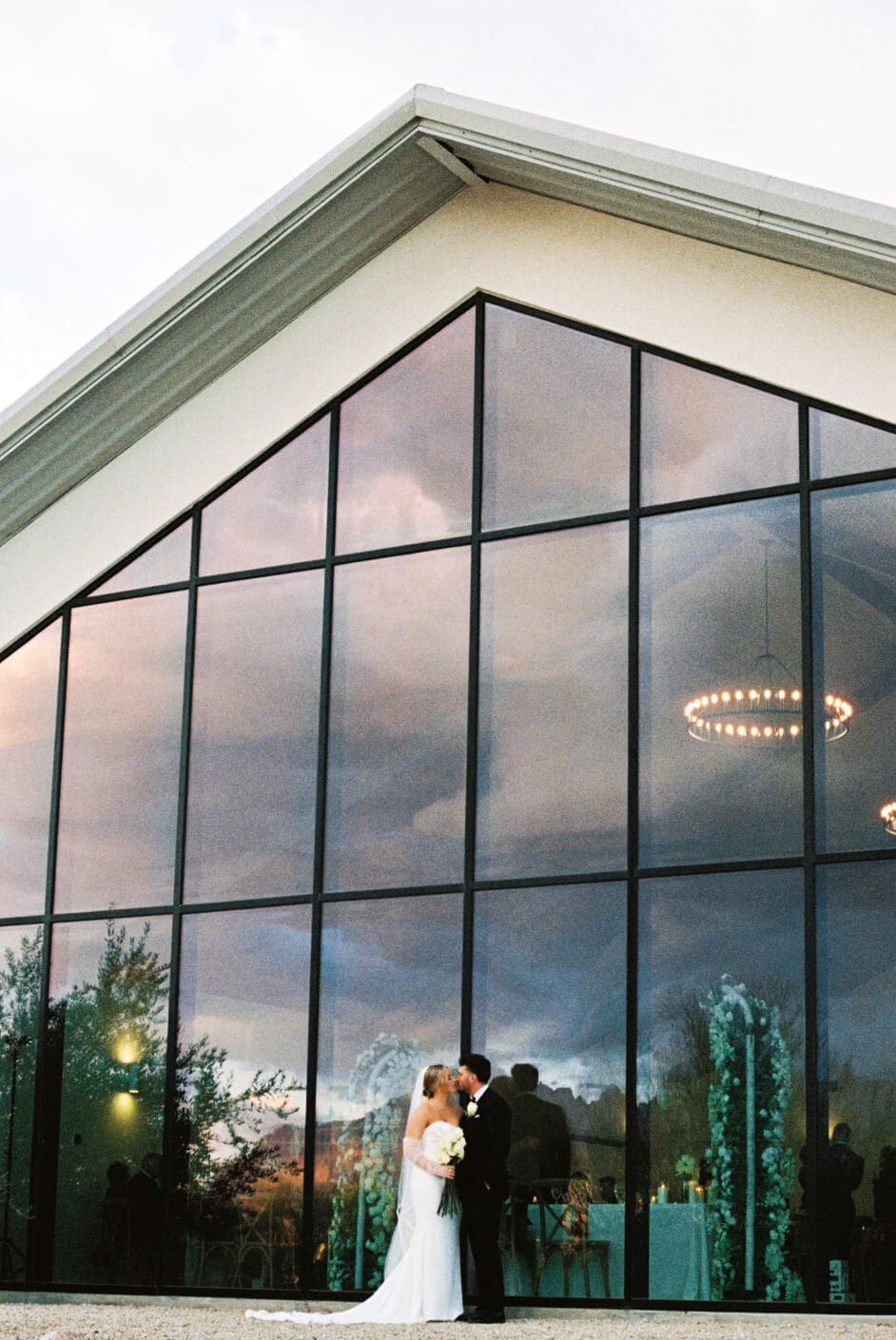 Bride and groom stand beneath the dramatic glass facade of a modern Arizona wedding venue at dusk.