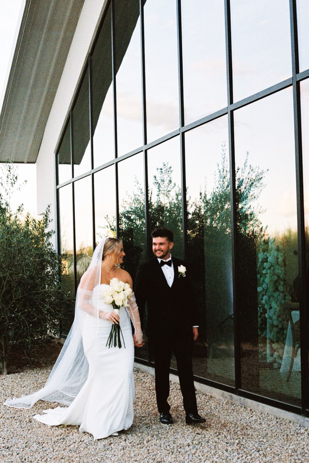 Bride and groom pose outside a modern glass wedding venue surrounded by desert landscaping and tall cacti.