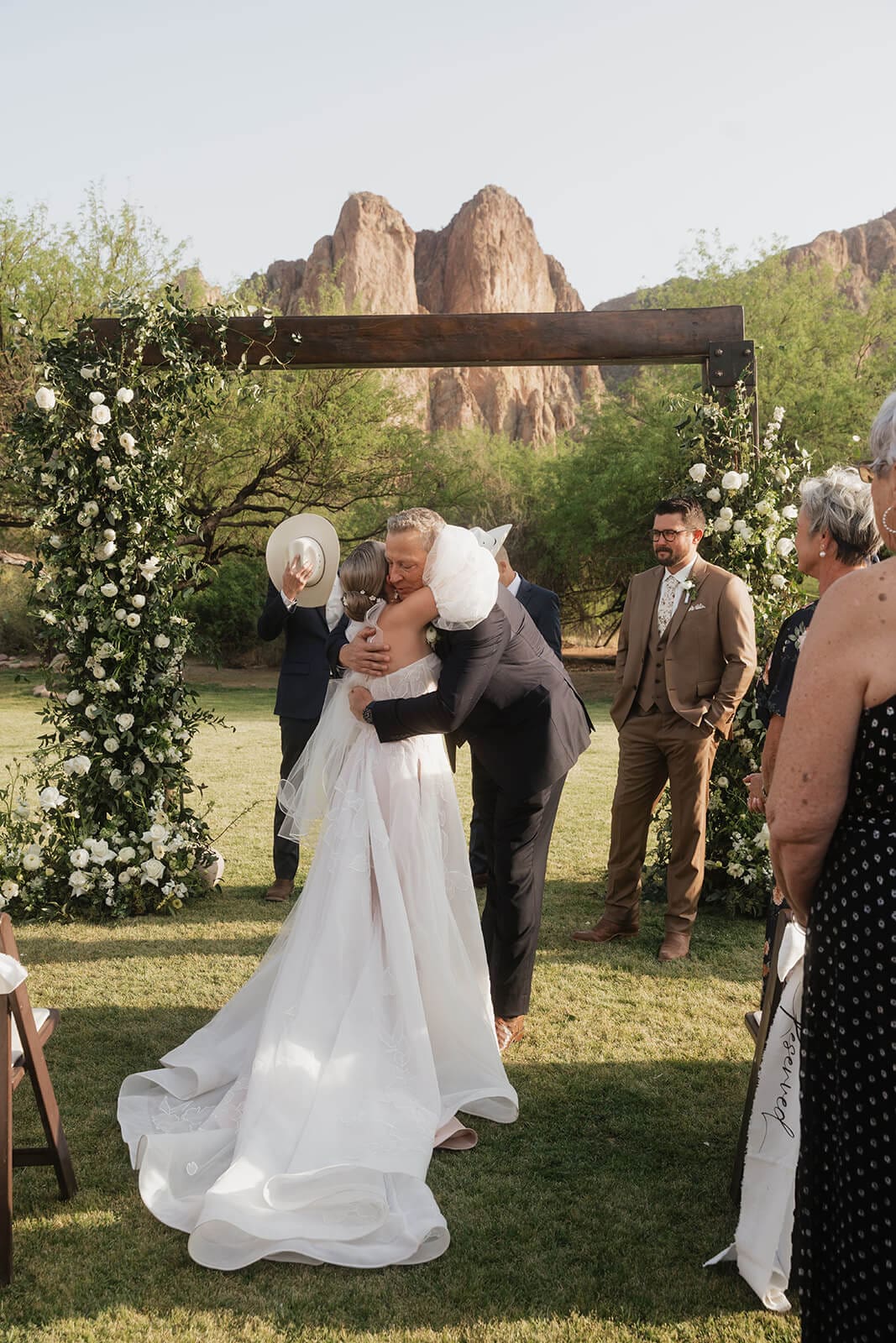 Bride embraces a family member during an outdoor wedding ceremony with the groom and Superstition Mountains in the background.