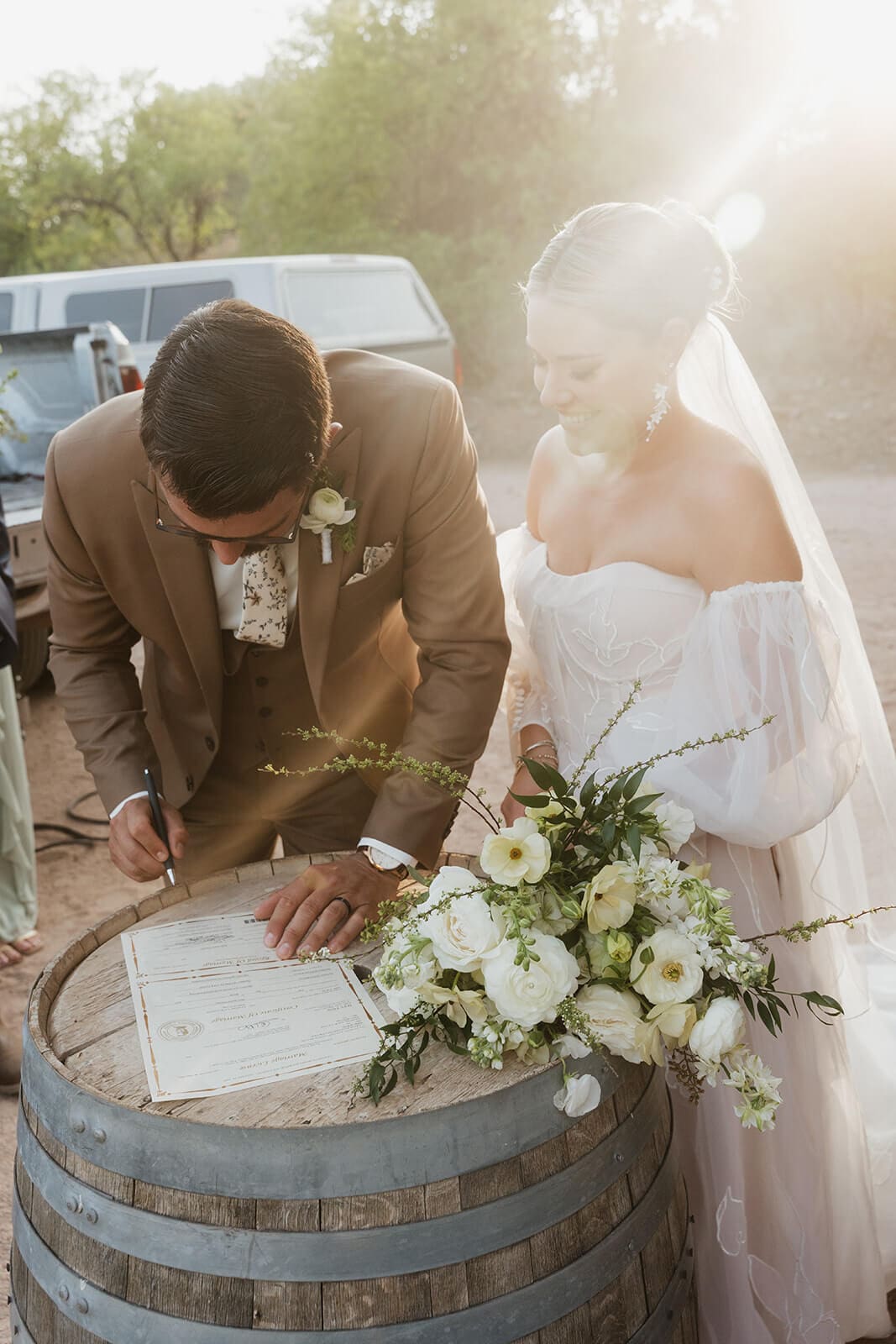 Bride and groom sign their marriage license on a wooden barrel as warm sunlight filters across the desert wedding venue.