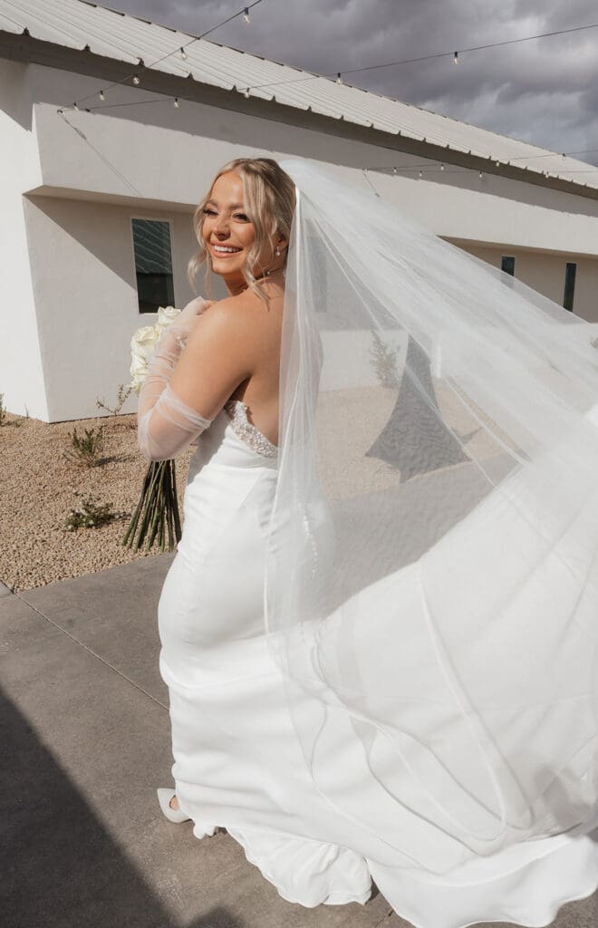 Bride looking over her shoulder outdoors as her long veil catches the light behind her.