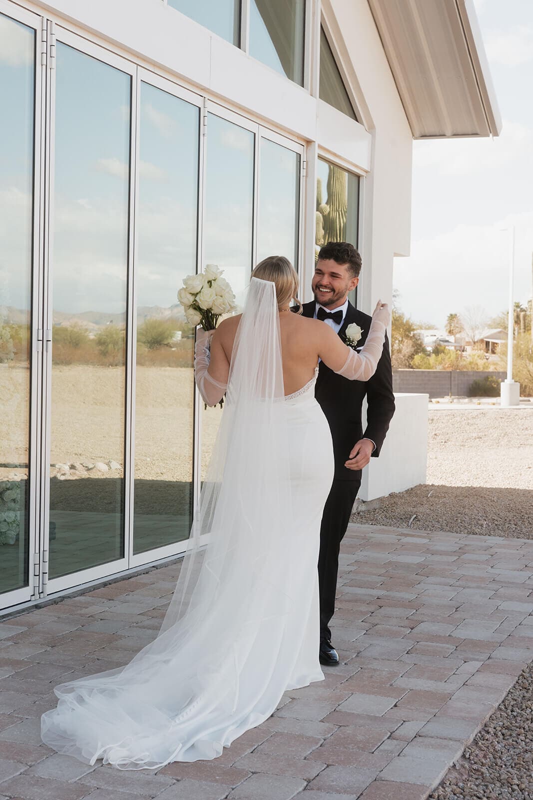 Bride approaches the smiling groom during their first look beside reflective glass windows and desert landscaping.