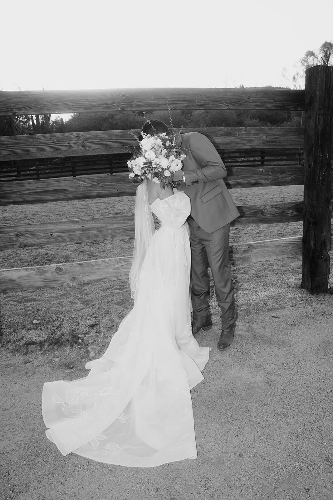 Black-and-white portrait of a bride and groom embracing beside a rustic wooden fence after their Arizona wedding ceremony.