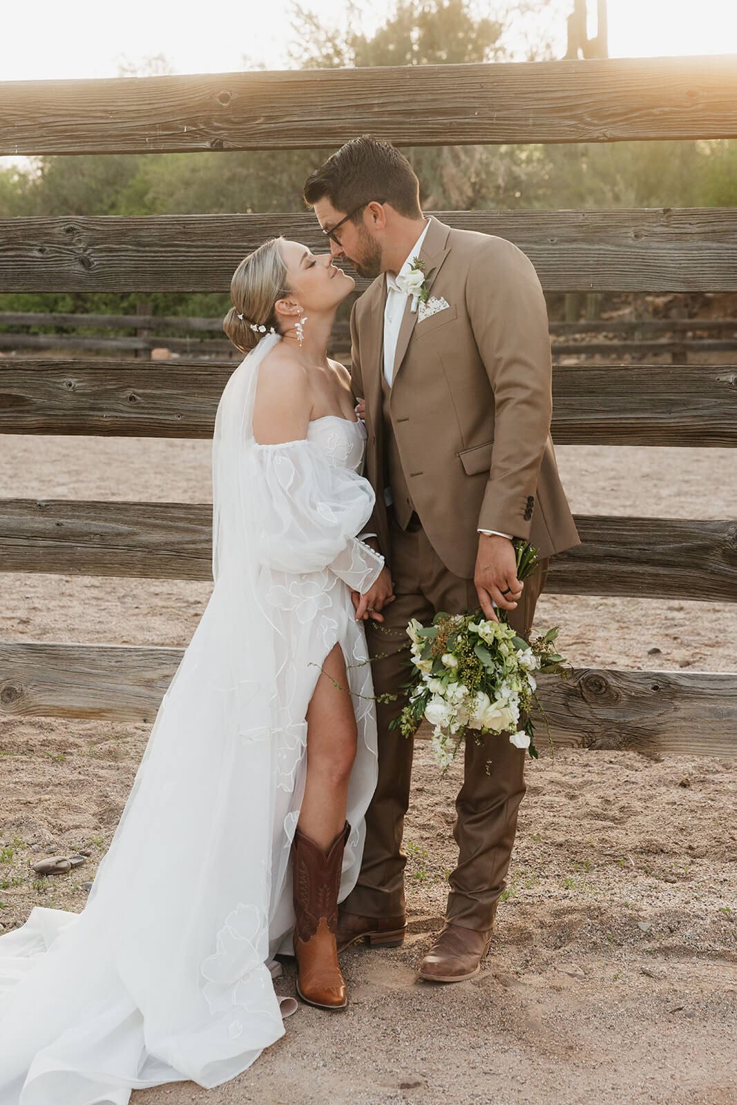 Bride in an off-the-shoulder wedding gown kisses the groom in a brown suit beside a rustic wooden fence.