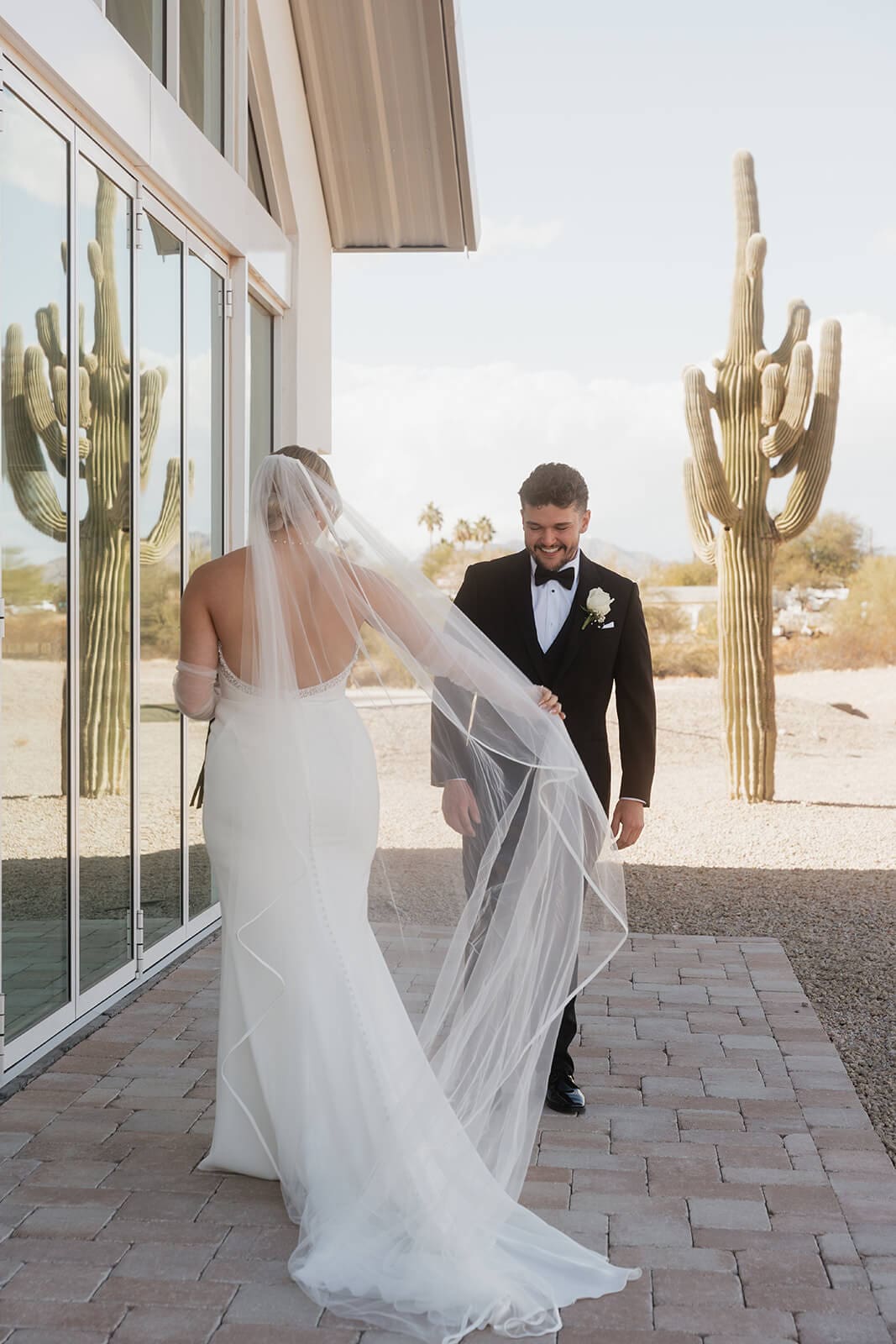 Bride walks toward the smiling groom outside their wedding venue as her long veil catches the desert breeze.