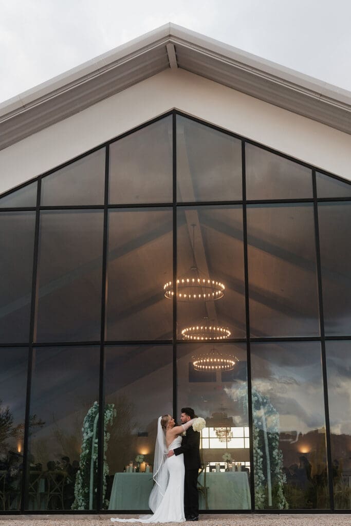 Newlyweds kissing in front of a modern glass chapel with glowing circular chandeliers visible inside.