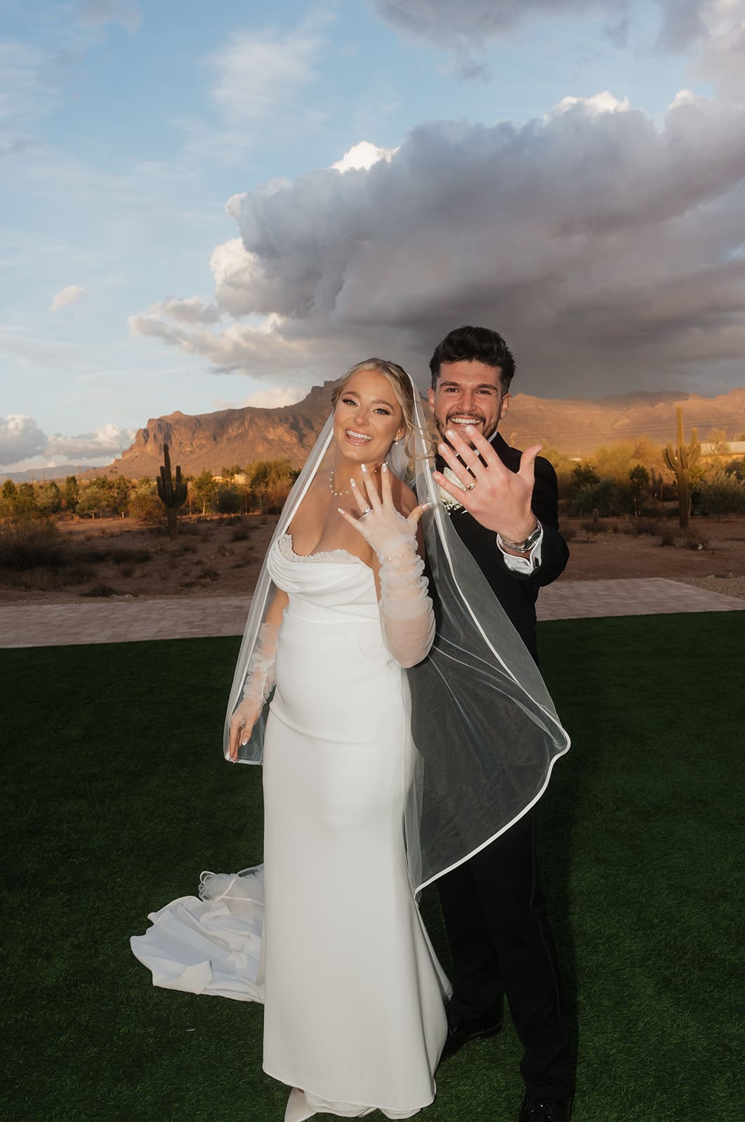 Newlyweds smile and show their wedding rings to the camera beneath dramatic clouds at sunset in the Arizona desert.