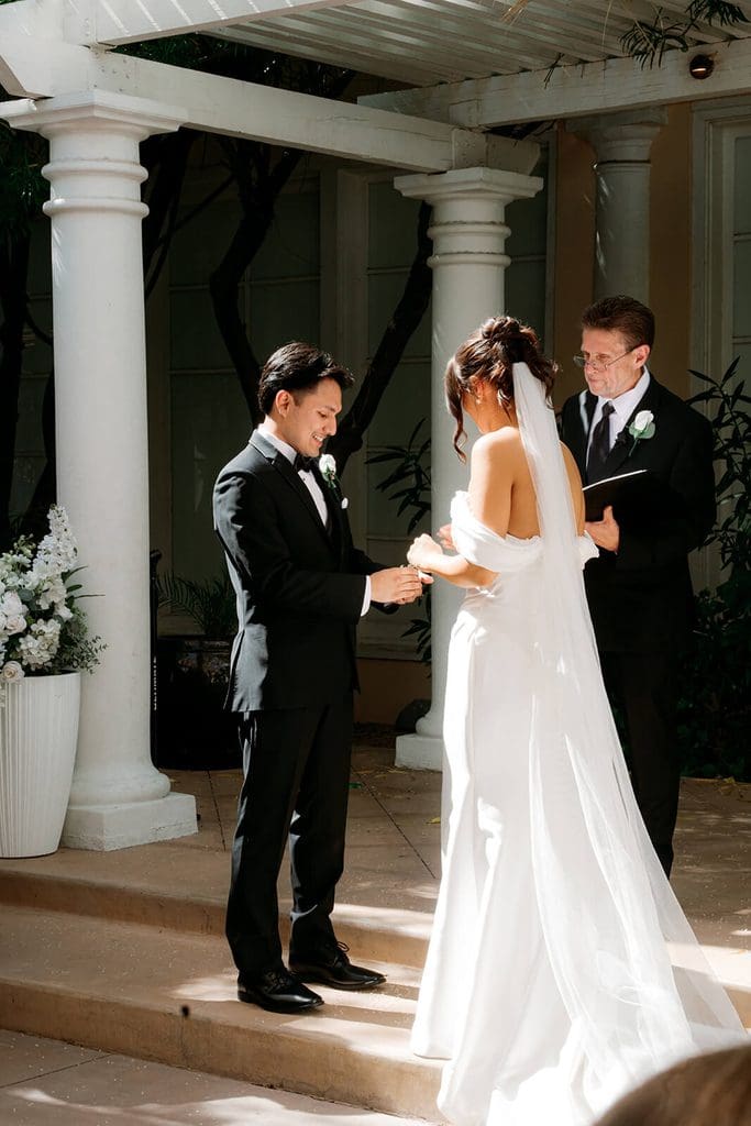 Bride and groom exchanging vows during an outdoor ceremony beneath a white pergola and columns.