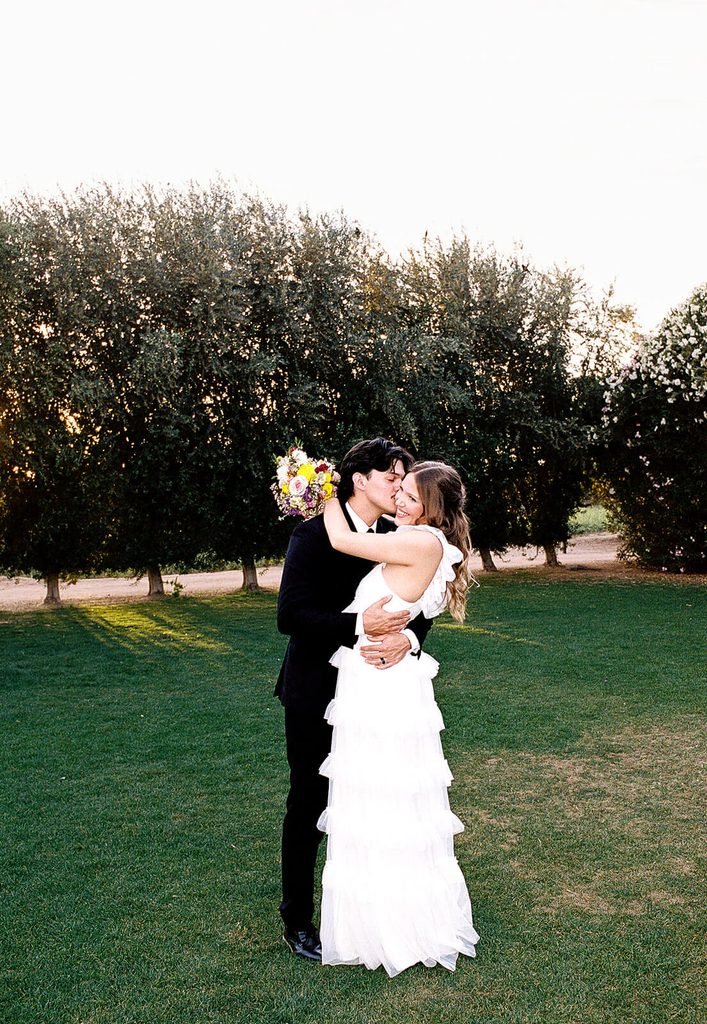 Bride and groom embracing on a green lawn with rows of olive trees in the background.