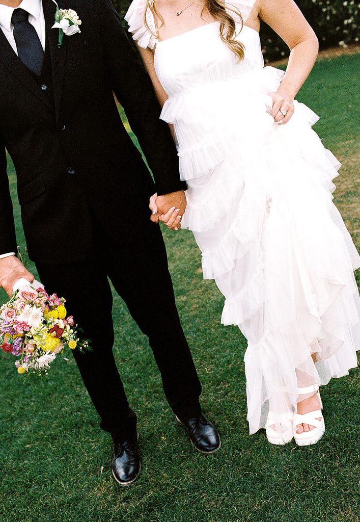 Cropped wedding portrait showing the couple holding hands, the bride’s tiered dress, and colorful bouquet.