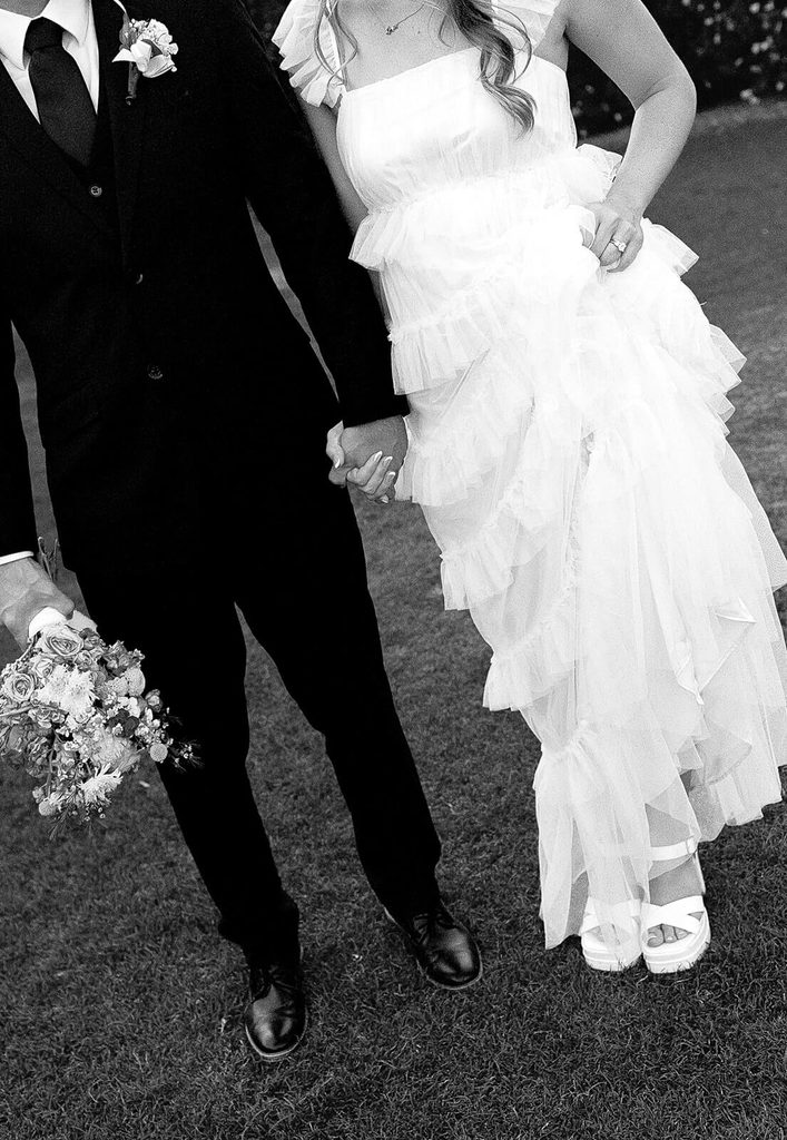 Black-and-white detail photo of the newlyweds holding hands and showing the bride’s tiered wedding dress.