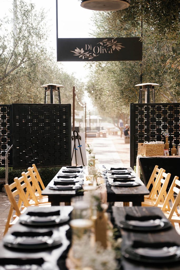 Outdoor wedding reception table set with black plates beneath olive trees and a Di Oliva sign.