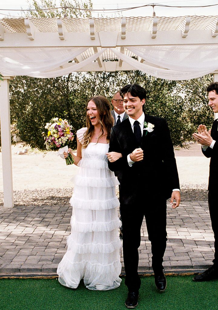 Bride and groom smiling as they walk away from their outdoor wedding ceremony beneath a white pergola.
