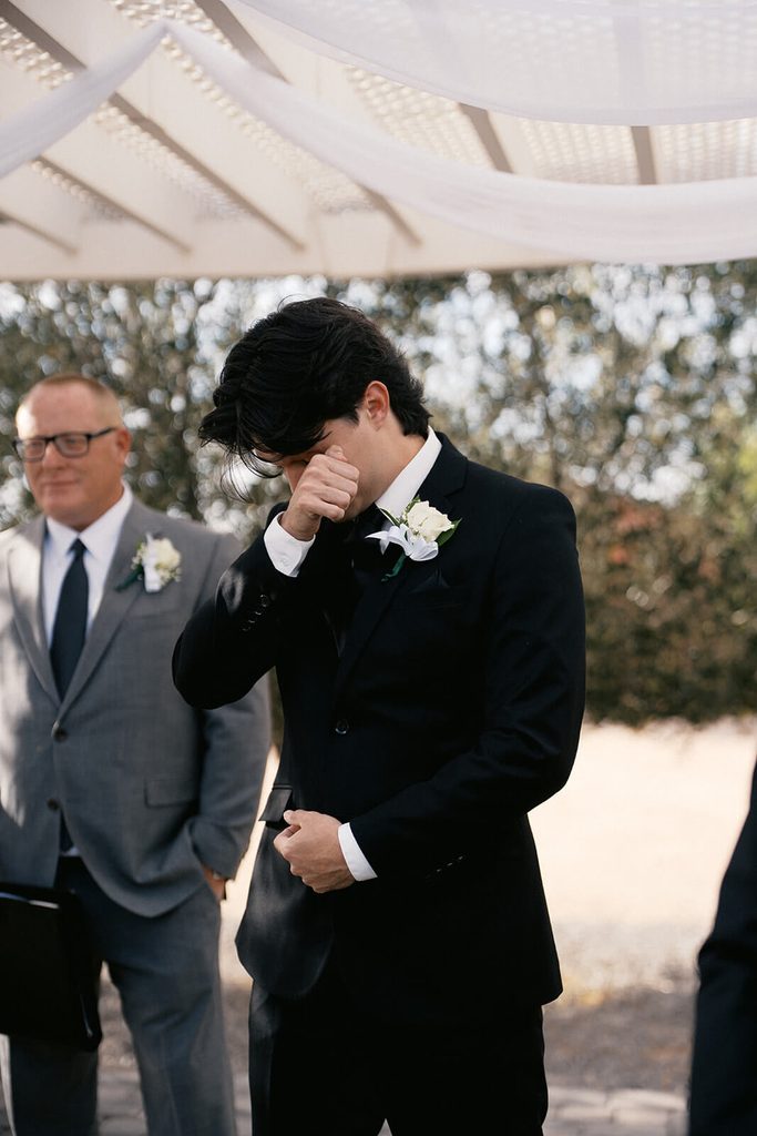 Emotional groom wiping away a tear during the wedding ceremony beneath a white pergola.