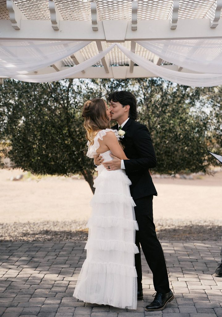 Newlyweds sharing their first kiss beneath a white pergola at their outdoor Arizona wedding.