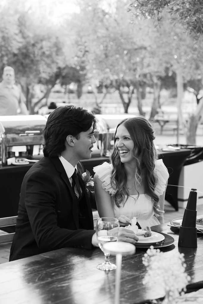 Black-and-white photo of newlyweds smiling together while seated at their reception table.