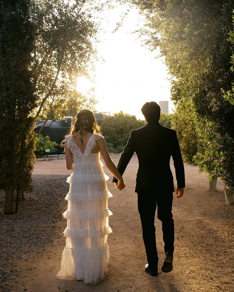 Bride and groom walking hand in hand through an olive grove with golden sunlight behind them.