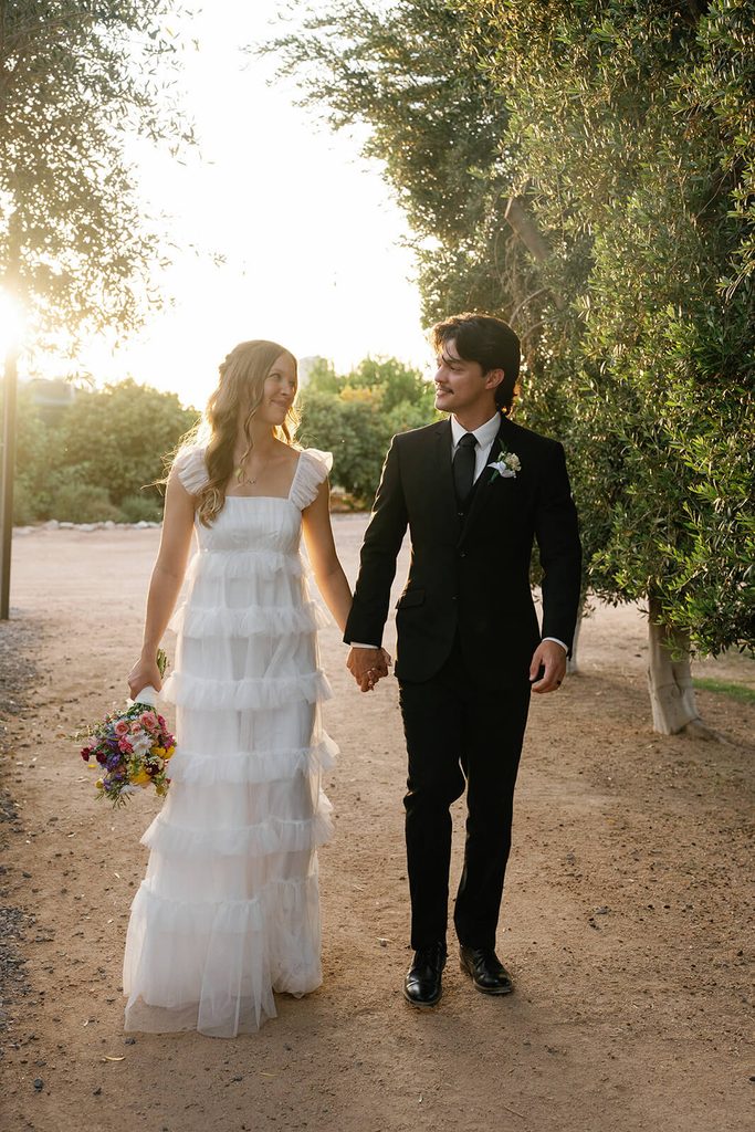 Bride and groom holding hands while walking through an olive grove in warm golden-hour light.