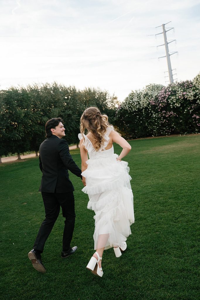 Bride and groom walking hand in hand across the lawn with olive trees behind them.