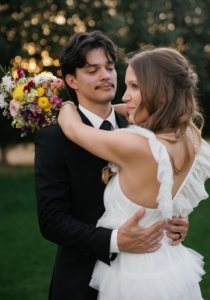 Newlyweds embracing on a green lawn with trees glowing in the background.