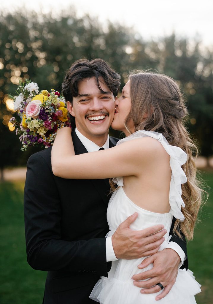Bride kissing the groom on the cheek as he smiles during outdoor wedding portraits.