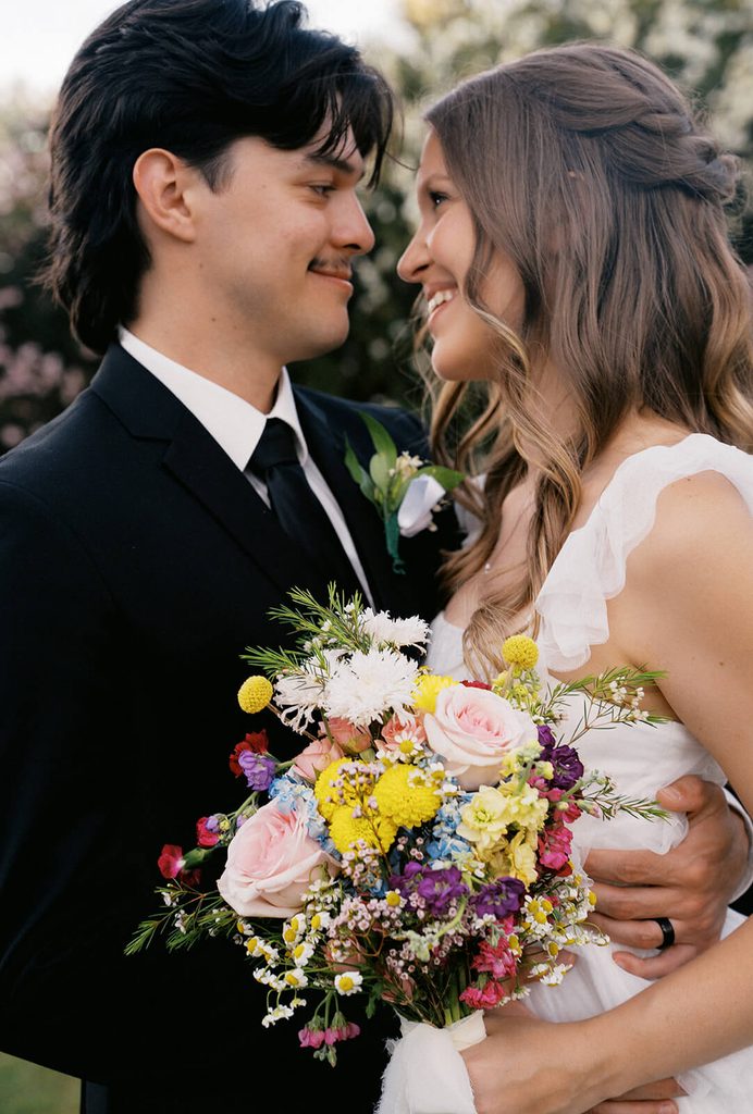 Close-up portrait of a bride and groom smiling at each other while holding a colorful wedding bouquet.