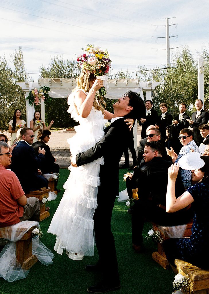 Groom lifting the bride into the air as guests celebrate around them after the ceremony.