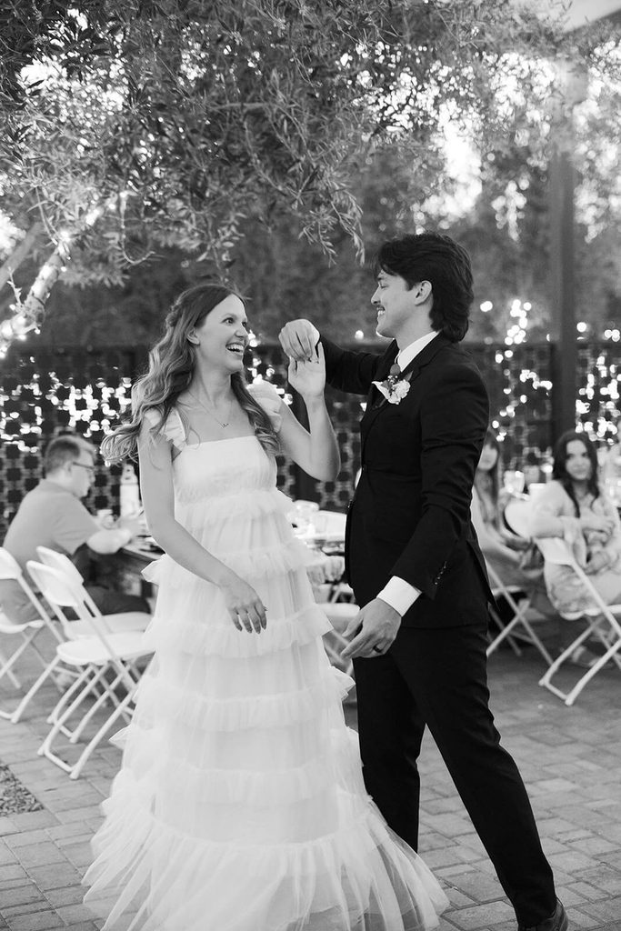 Black-and-white photo of the groom twirling the bride beneath string lights during their outdoor reception.