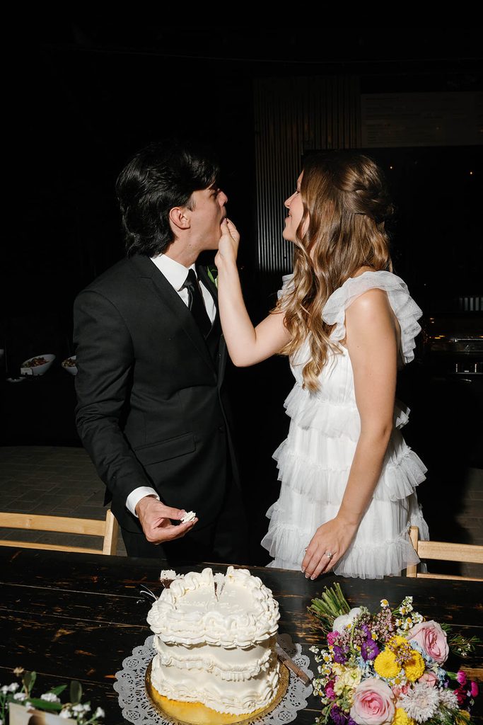 Bride playfully feeding the groom cake beside their white wedding cake and colorful bouquet.