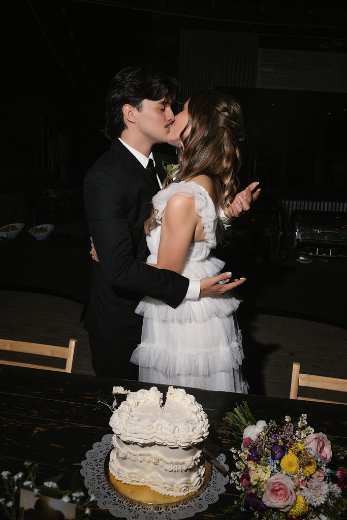 Newlyweds kissing beside their vintage white wedding cake during the reception.
