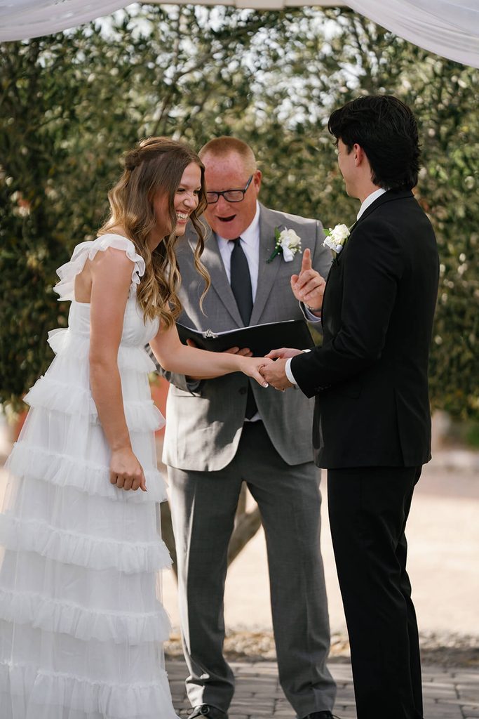 Bride and groom laughing together during their outdoor wedding ceremony with their officiant behind them.