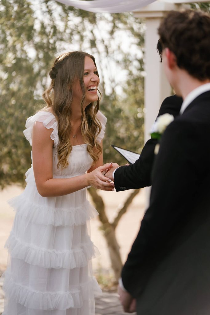 Bride laughing as she holds the groom’s hand during their outdoor wedding ceremony.
