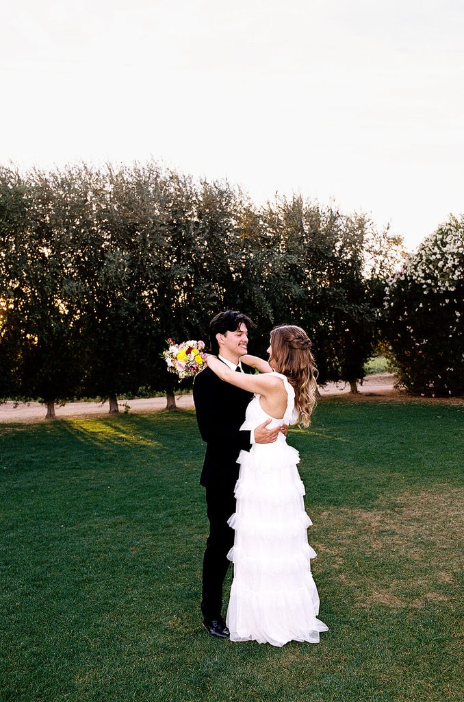 Newlyweds embracing on a green lawn surrounded by olive trees at Queen Creek Olive Mill.